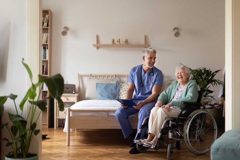 A caregiver assists an older woman with a gentle walk in a residential hallway decorated with personal photos.